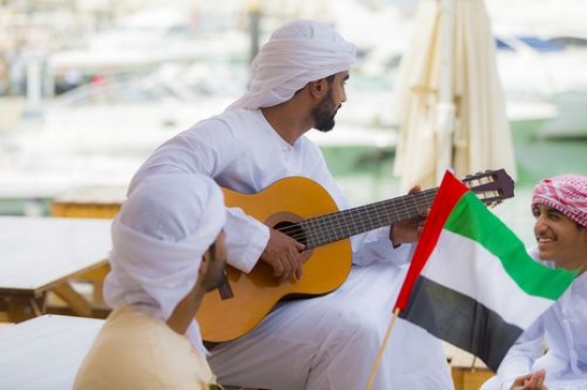Arab man carrying a UAE Flag while the another man is playing an instrument|