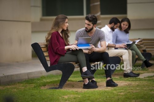 University Students sitting on the bench talking to each other while using the device|-