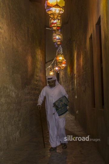 Arab man carrying a prayer rug while walking through the alleyway of Ramadan Lanterns|-