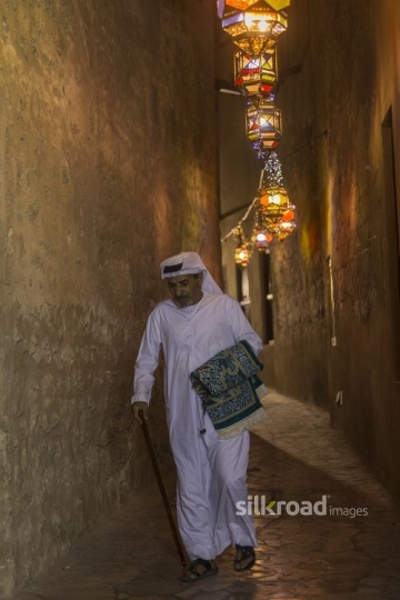 Middle Eastern man carrying a prayer rug while walking through the pathways of Ramadan Lanterns|-